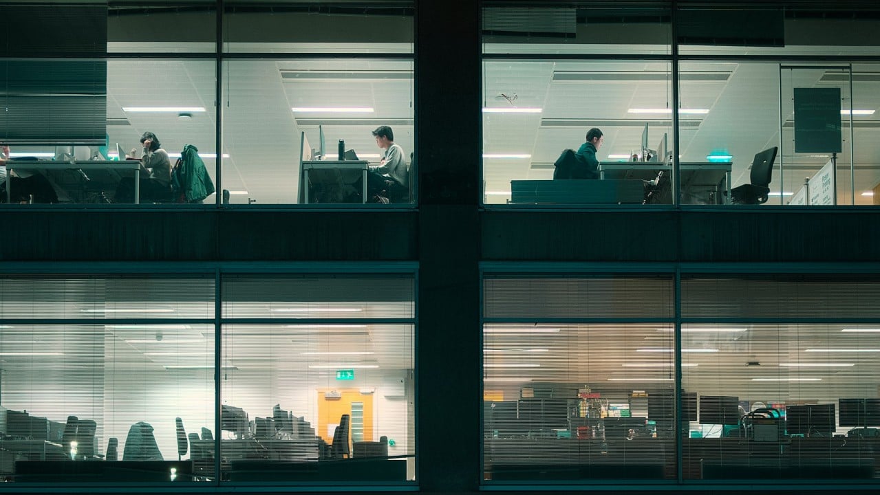 Office building exterior at dusk showing illuminated windows with silhouettes of workers at desks across multiple floors