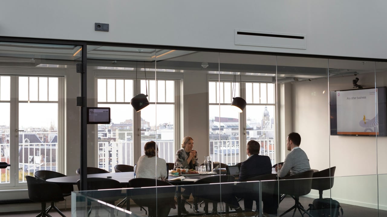 Five people in business meeting around a table in modern office with floor-to-ceiling windows overlooking a city skyline